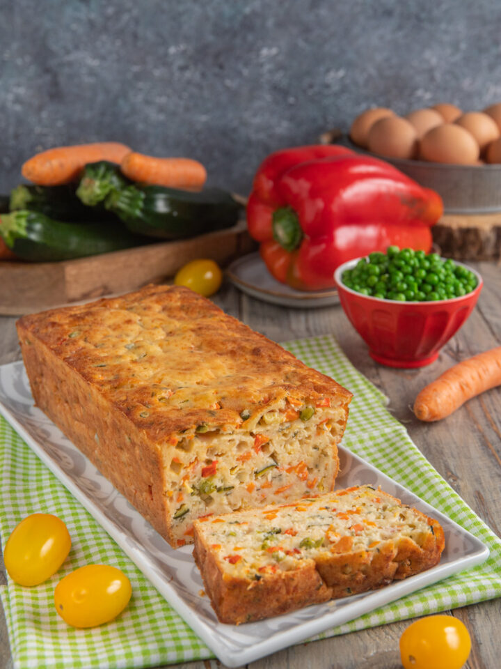 Torta salata con yogurt e verdure su piatto rettangolare, con una fetta tagliata, pomodorini gialli, ciotolina di piselli e peperone rosso sullo sfondo