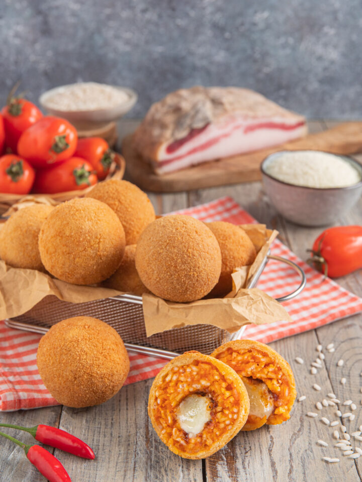 Arancini all'amatriciana in contenitore di alluminio con carta paglia, su tovaglietta a quadretti bianchi e rossi e arancino diviso a metà in primo piano