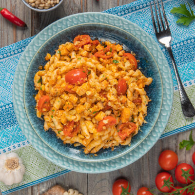 Busiate al ragù di pesce spada impiattate in piatto azzurro, su tovaglietta azzurra con attorno pomodorini, peperoncino, aglio e forchetta argentata sulla destra