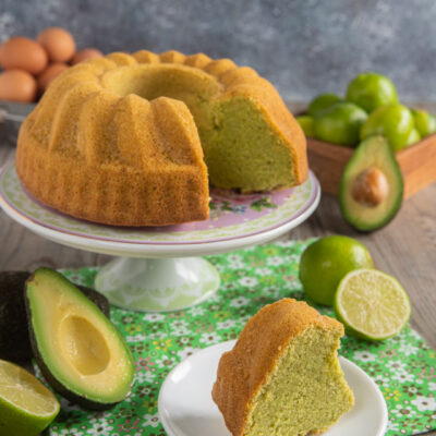 Ciambella all'avocado su alzatina bianca, con fetta tagliata, tovaglietta verde e avocadi e lime tagliati