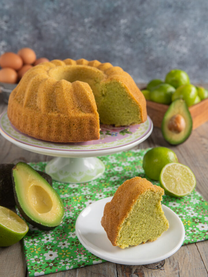 Ciambella all'avocado su alzatina bianca, con fetta tagliata, tovaglietta verde e avocadi e lime tagliati