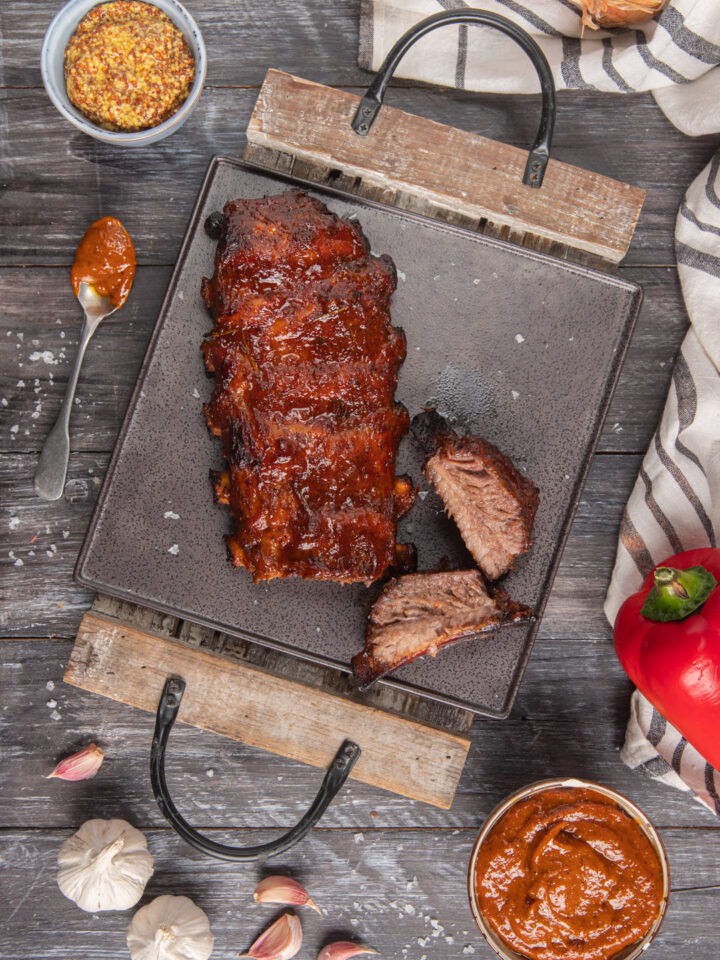 Costine di maiale in salsa bbq su vassoio quadrato di metallo e legno, con manici, peperone rosso e ciotolina con salsa bbq