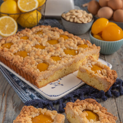 Crumble cake alle pesche con tre cubotti tagliati, limoni sullo sfondo, pesche sciroppate e mandorle accanto