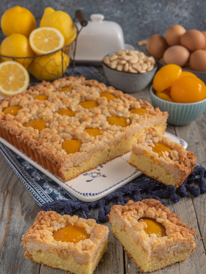 Crumble cake alle pesche con tre cubotti tagliati, limoni sullo sfondo, pesche sciroppate e mandorle accanto