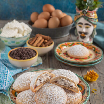 Spagnolette siciliane su piattino tondo, intere più una spezzata. Sullo sfondo ciotoline con gocce di cioccolato, cannella in stecche, ricotta, uova e statuina di testa di moro