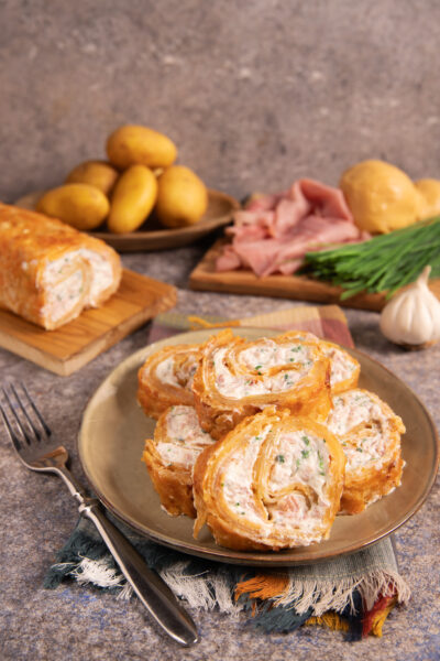 Rotolo di patate croccante con patate sottili e dorate al forno, avvolge un morbido ripieno di formaggi e prosciutto cotto.