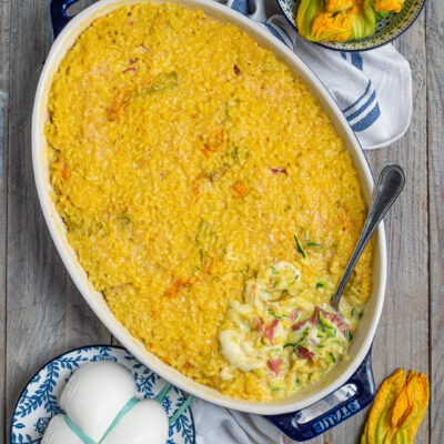 Il riso al forno con zucchine, fiori di zucca, speck e mozzarella è una gustosa variante del classico risotto, che si compone a strati e termina la cottura in forno per formare una bella crosticina; l'ideale per un pranzo all'insegna della convivialità.