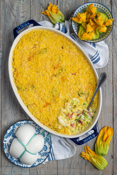Il riso al forno con zucchine, fiori di zucca, speck e mozzarella è una gustosa variante del classico risotto, che si compone a strati e termina la cottura in forno per formare una bella crosticina; l'ideale per un pranzo all'insegna della convivialità.