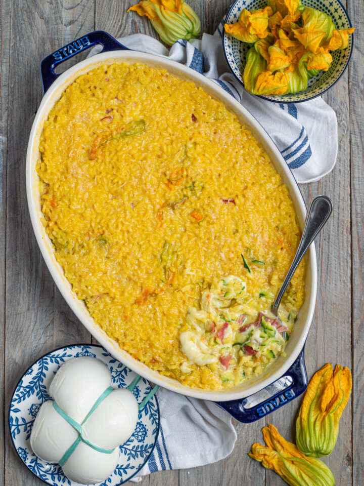 Il riso al forno con zucchine, fiori di zucca, speck e mozzarella è una gustosa variante del classico risotto, che si compone a strati e termina la cottura in forno per formare una bella crosticina; l'ideale per un pranzo all'insegna della convivialità.